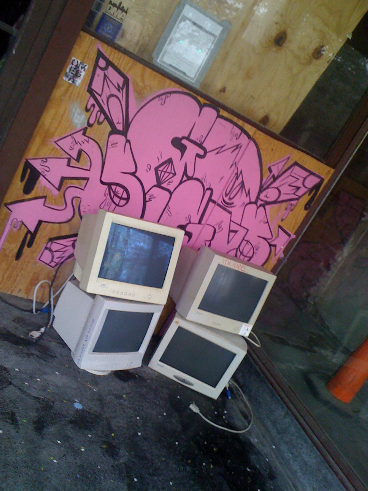 Four CRT computer monitors stacked in front of an outdoor wall covered with grafiti.
