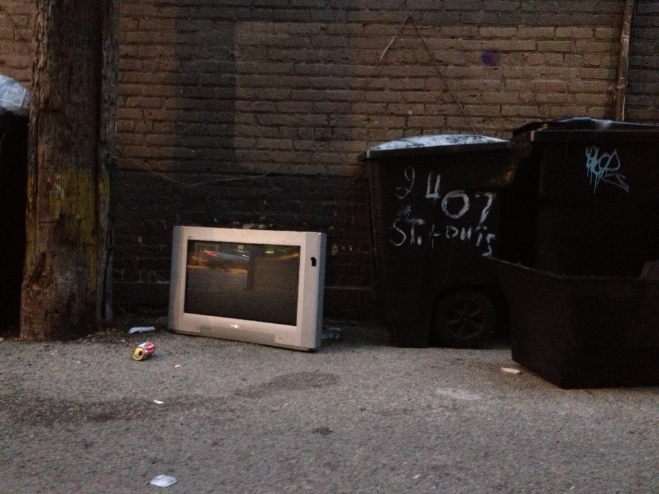 A CRT television photographed in an alley near two trash bins.