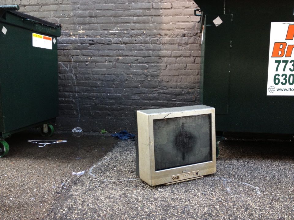 A CRT television photographed in an alley near two dumpsters. The screen has a black circle in the middle from being broken.