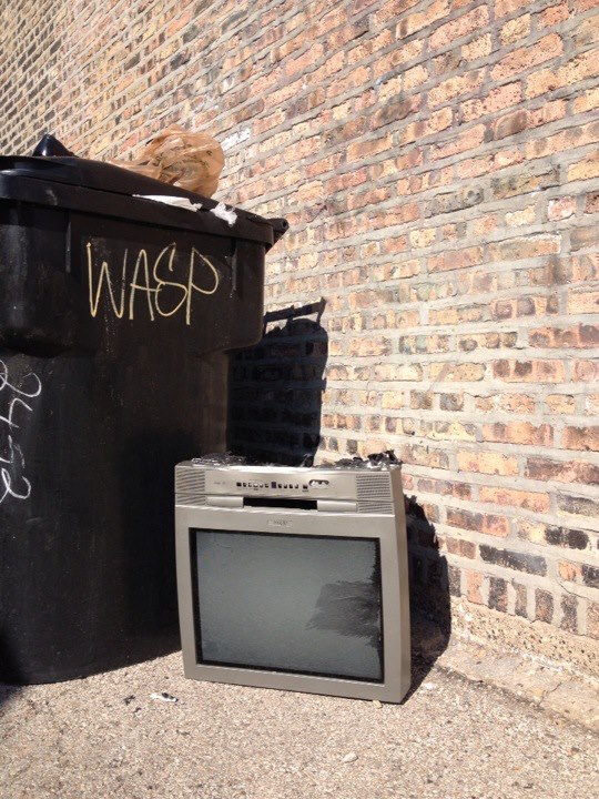 An upside down cathode way tube with the bottom smashed open, photographed in an alley beside a dumpster.