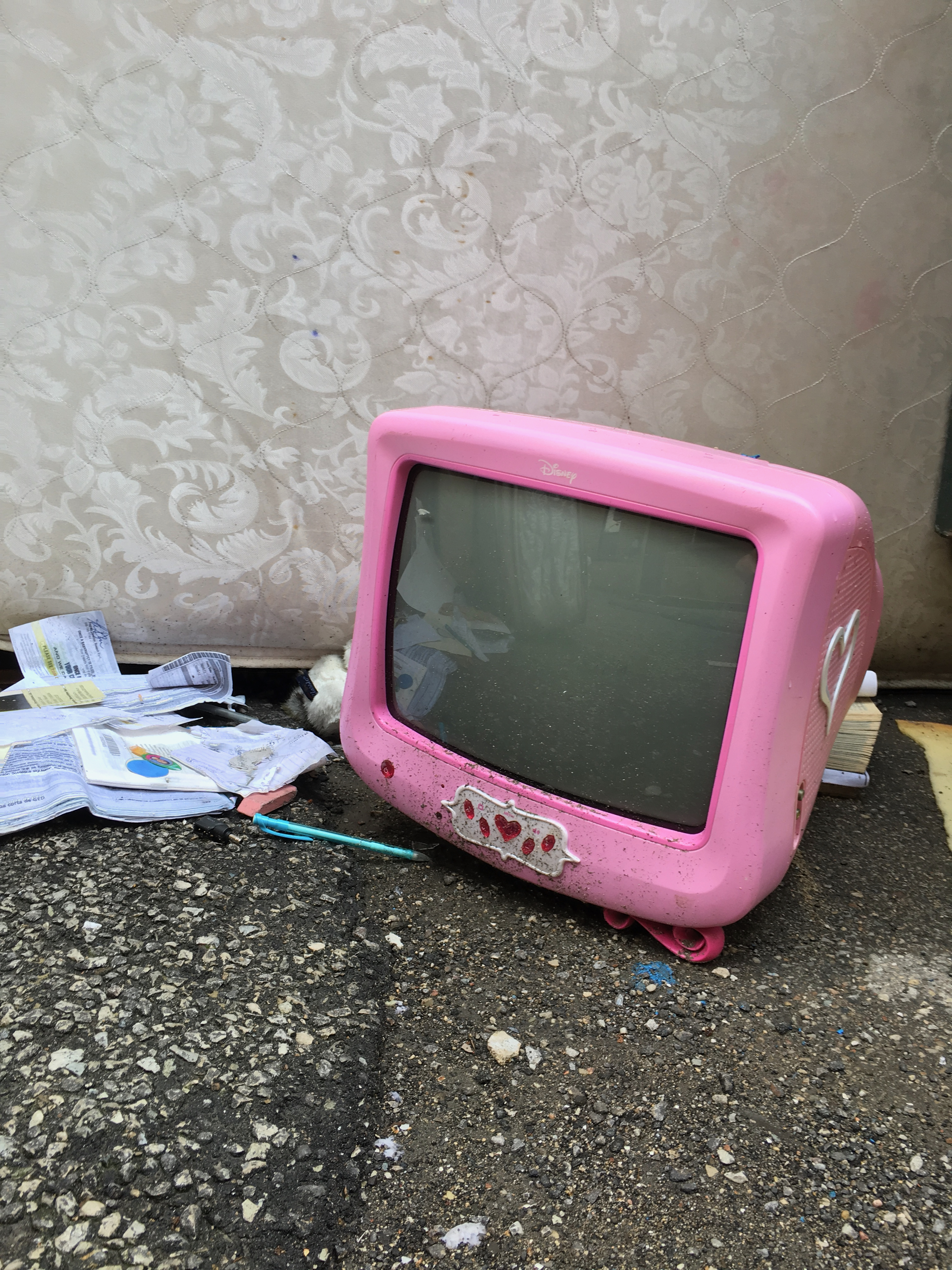 A small pink CRT television with a white heart on the side and jewel buttons photographed in an alley with a couch in the background and discarded school supplies next to it..