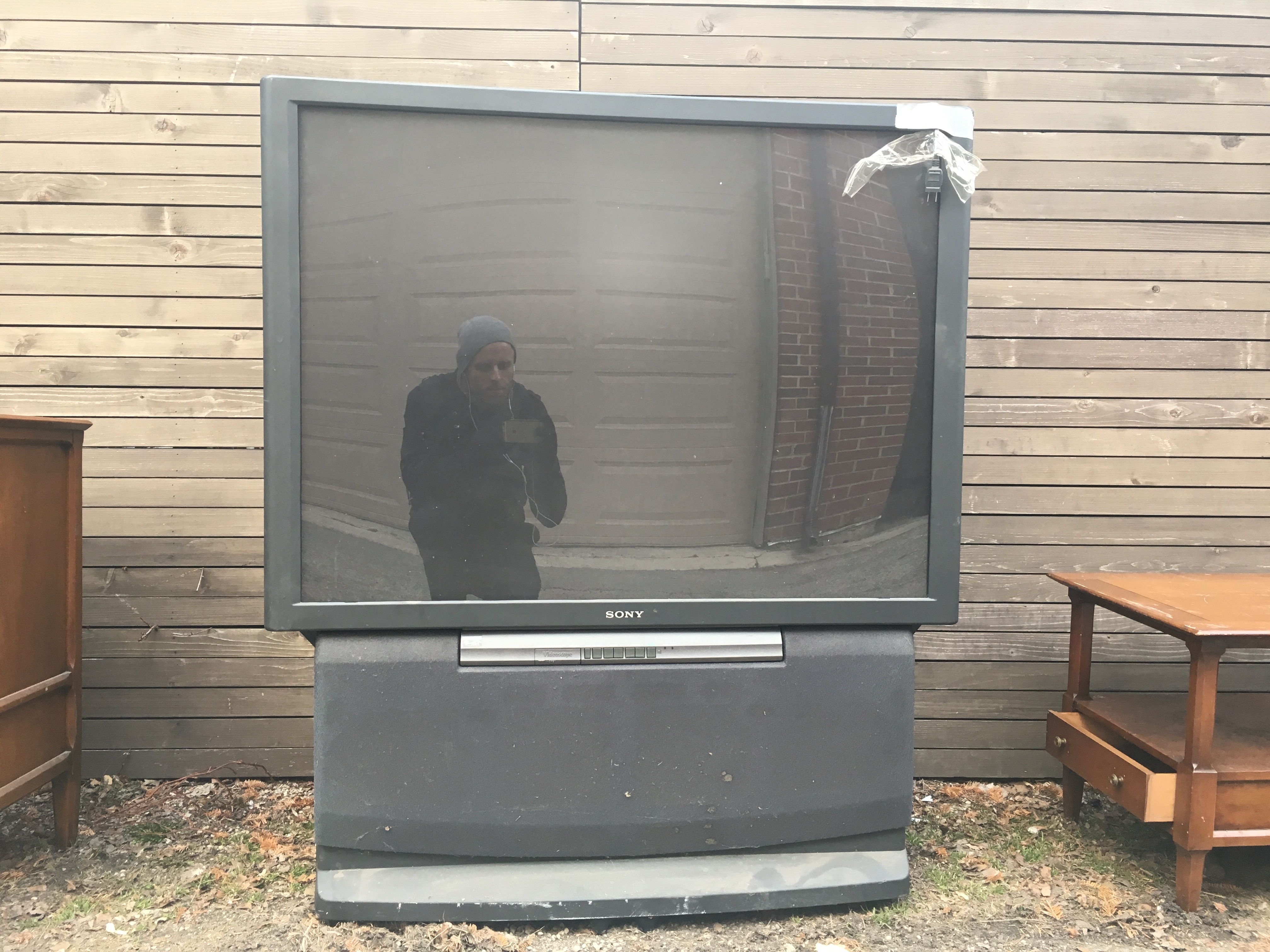 A large CRT televisipon photographed in an alley between two living room end tables, with the photographer reflected in the screen.