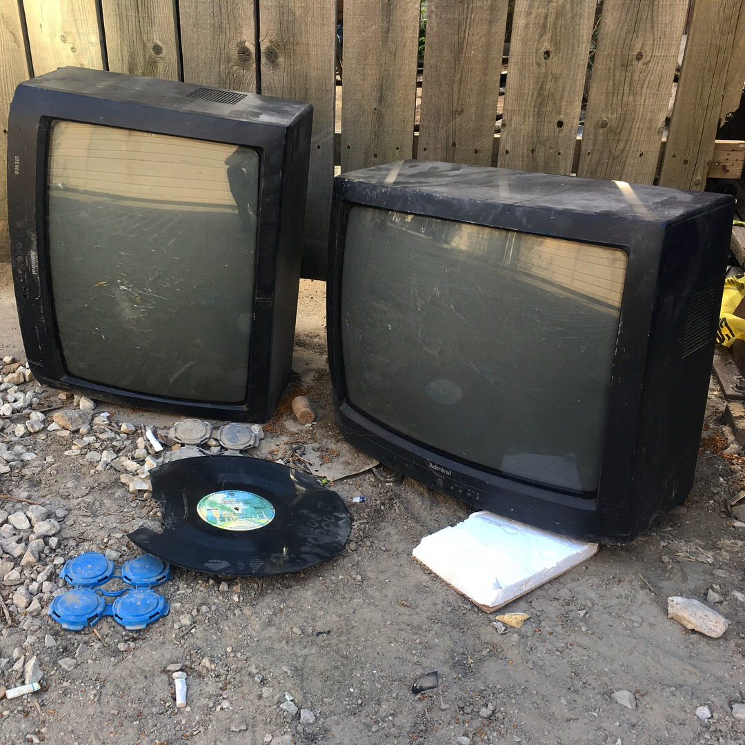 Two small CRTs on the ground near a wooden fence with a broken vinyl record in front of them.