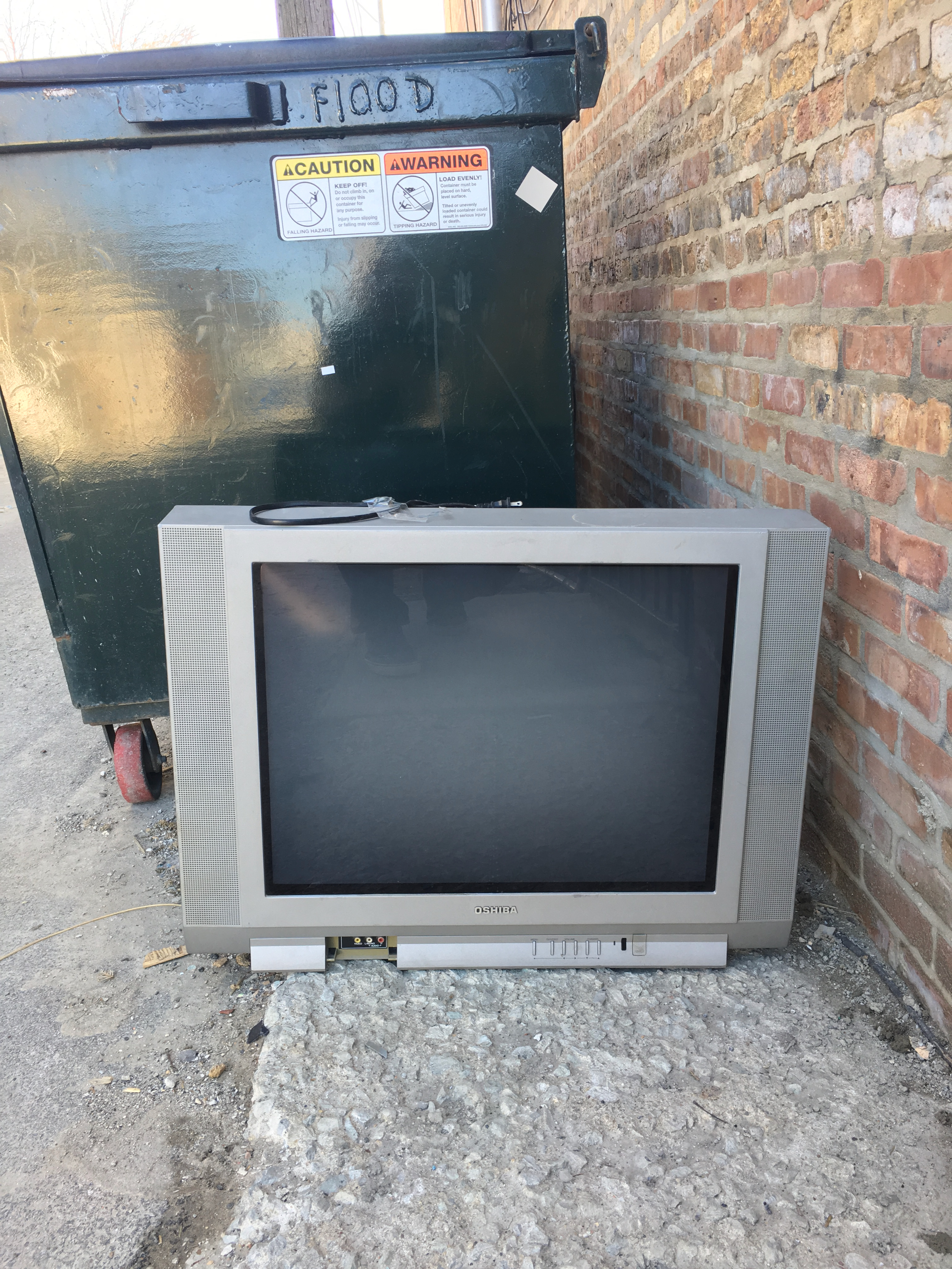 Large CRT television in photographed beside a dumpster in an alley.