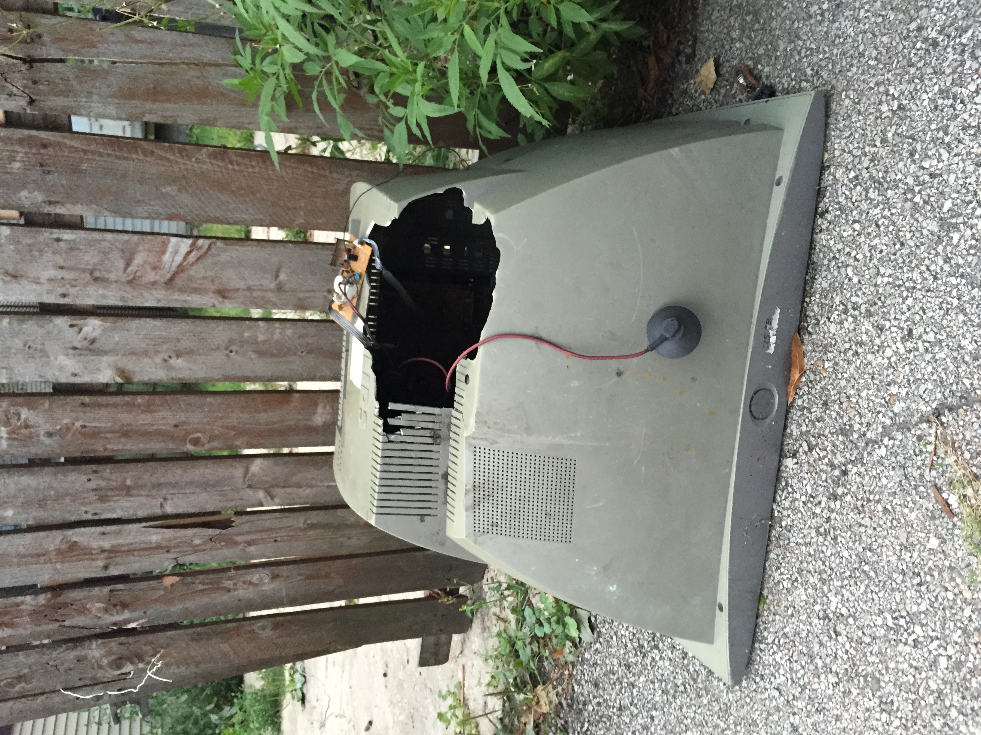 A large cathode ray tube television photographed near a wooden fence face down with its plastic case broken and a wire and suction cup coming out of the jagged hole.