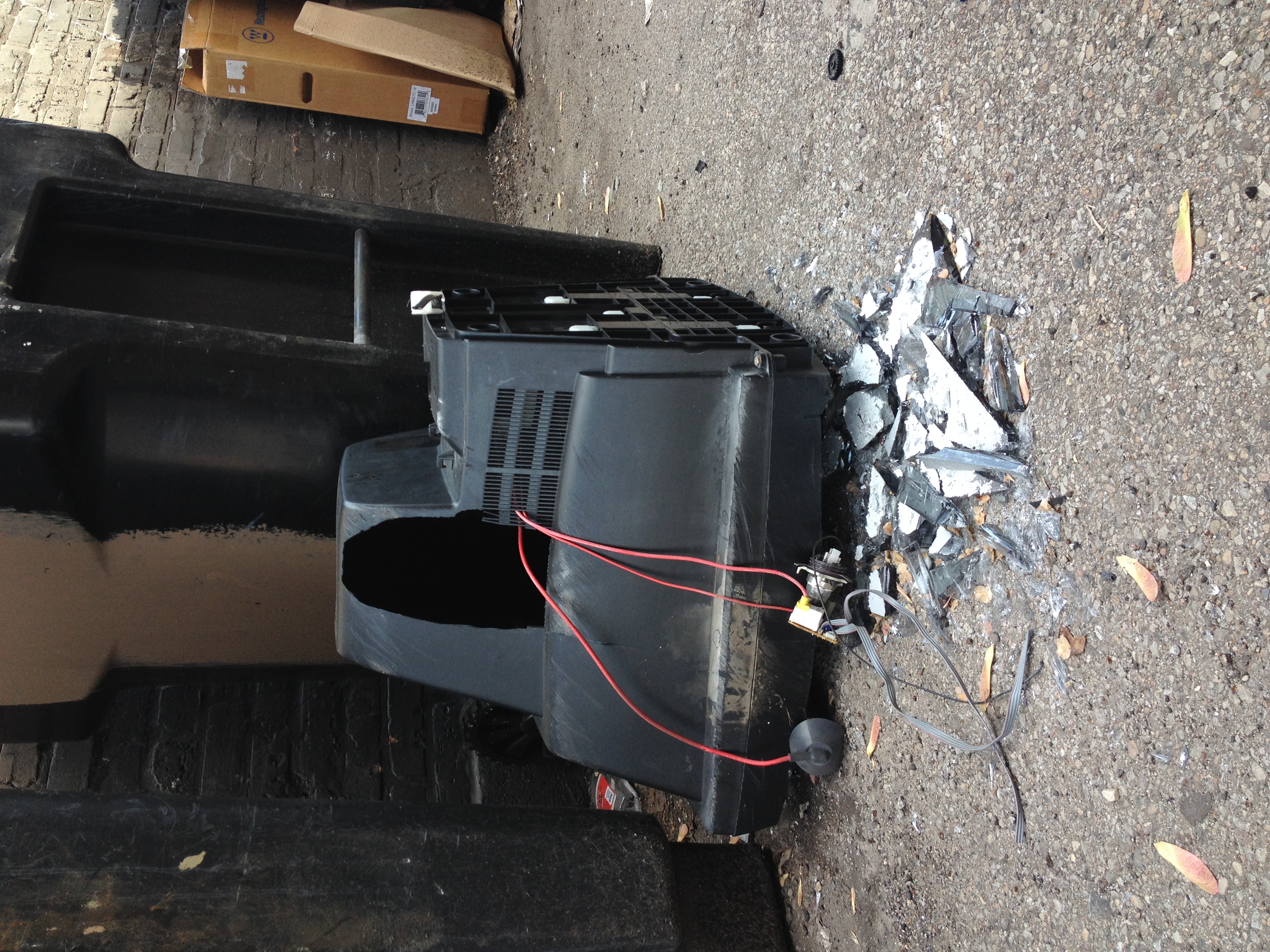 A CRT television photographed in an alley face down and broken with pieces on glass on the sement and wires spilling out of a hole in the plastic casing.